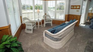A cozy room with a wicker sofa, two chairs, a small table, and a lamp. Large windows with blinds offer a view of the outside. - Earnest, Ships Watch Community in Duck, NC (Duck)
