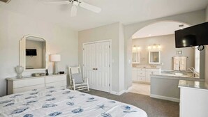 A bedroom with a bed, a chair, a desk, a television, and a bathroom with a sink and mirrors.