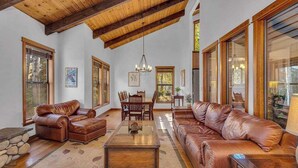 A spacious living room with leather sofas, a wooden coffee table, and a dining area with a wooden table and chairs. Large windows allow natural light to fill the space. - Hot tub, large home, family friendly, ping pong, a premier Sunriver Retreat (Sunriver)