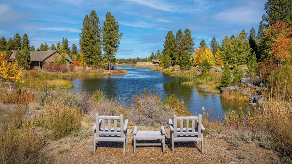 Vista al lago con dos sillas de madera orientadas al agua, rodeado de árboles y una cabaña.