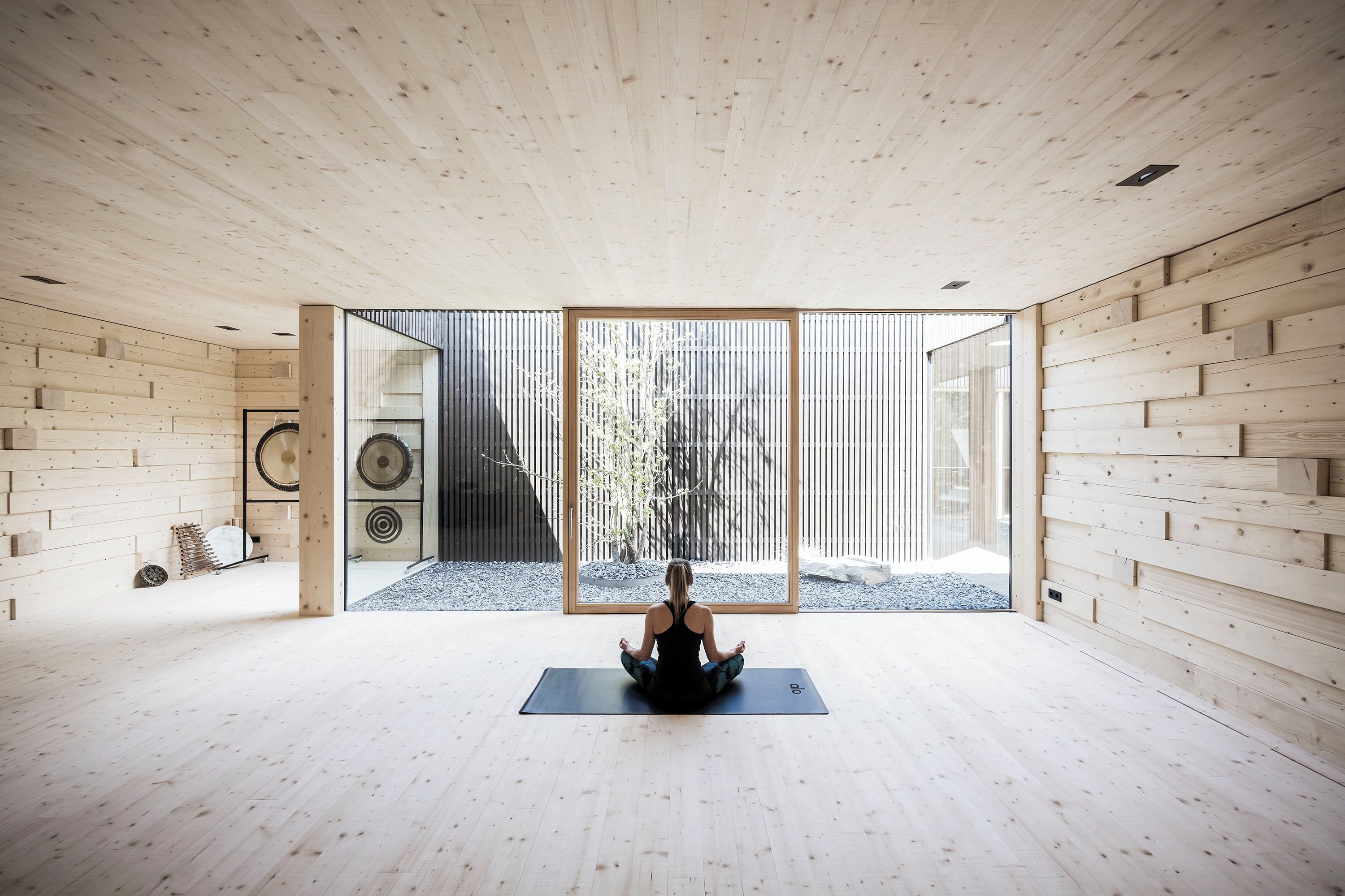Una habitación amplia con piso y paredes de madera, un gran ventanal corredero que da a un espacio exterior y una persona sentada sobre una colchoneta de yoga.
