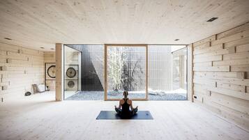Una habitación amplia con piso y paredes de madera, un gran ventanal corredero que da a un espacio exterior y una persona sentada sobre una colchoneta de yoga.