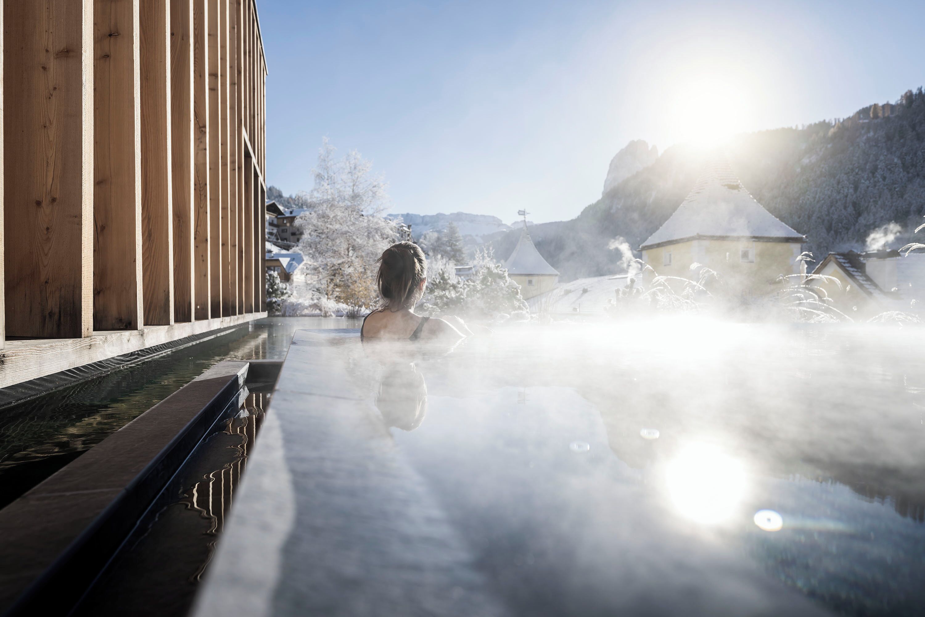A person in a hot spring with steam rising, surrounded by snow-covered buildings and a clear sky.