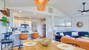 A modern kitchen with white cabinets, a microwave, and a refrigerator. There is a dining area with a wooden table and chairs, and a living room with a blue sofa and a ceiling fan. - Welcome to The Palms @ Sneads Ferry, NC! (Sneads Ferry)