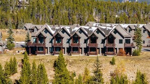 A row of log cabin-style buildings with balconies, set against a backdrop of snow-capped mountains and dense forests. - Saddle Ridge F5 3 Bedroom Home (Gallatin Gateway)