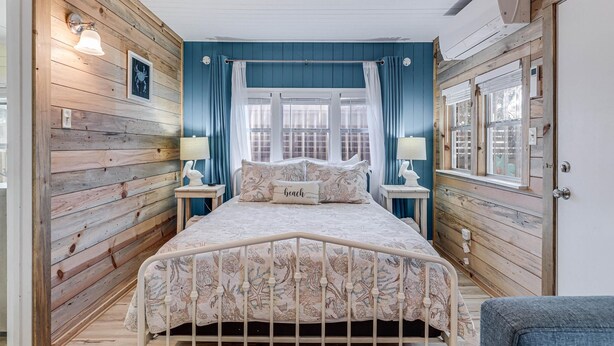 A bedroom with a wooden floor, a bed with a white frame, two bedside tables with lamps, a window with curtains, and a wall clock.