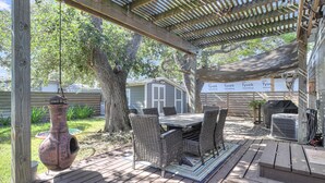A covered outdoor patio with a wooden table and wicker chairs, a large tree, and a grill.