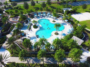 Eine Vogelperspektive zeigt einen Resortkomplex mit einem zentralen Schwimmbecken, umgeben von Palmen und Liegestühlen.