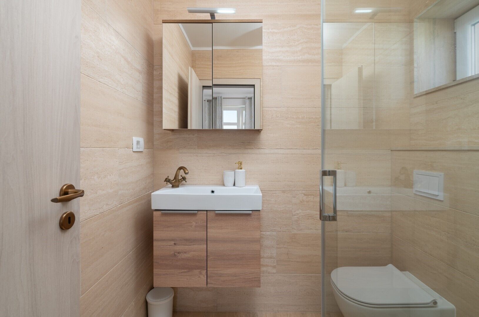 A modern bathroom with a glass shower enclosure, a white sink with a gold faucet, and wooden cabinets.
