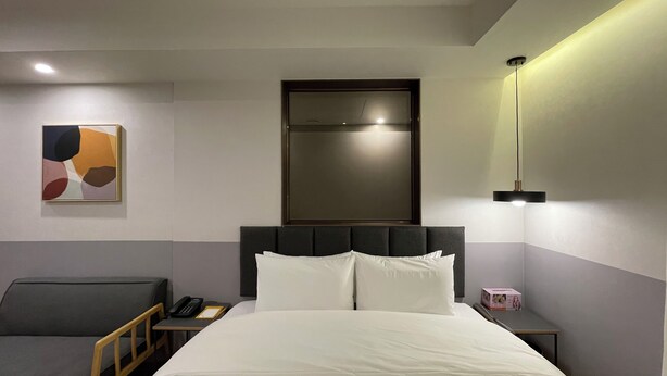 A hotel room with a bed, bedside table, chair, and a painting on the wall.