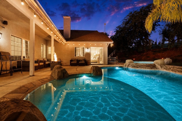 A swimming pool with a stone border, outdoor seating, and a building with a chimney.