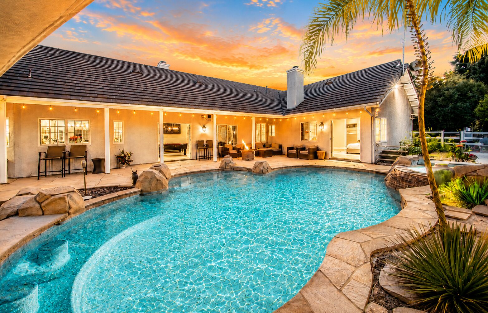 A swimming pool with a stone border, a covered patio area with seating, and a building with a tiled roof.