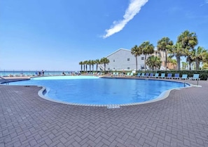 Una piscina con sillas de descanso y palmeras, un edificio de techo plano y un cielo despejado.