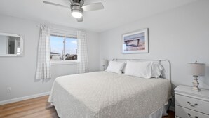 Une chambre à coucher comprenant un lit, une table de chevet, un ventilateur de plafond, une fenêtre avec des rideaux, un miroir fixé au mur et un tableau encadré.