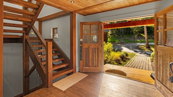 Un escalier en bois mène à une porte dotée d’un panneau de verre, le sol est en bois et on peut admirer un jardin depuis l’intérieur.