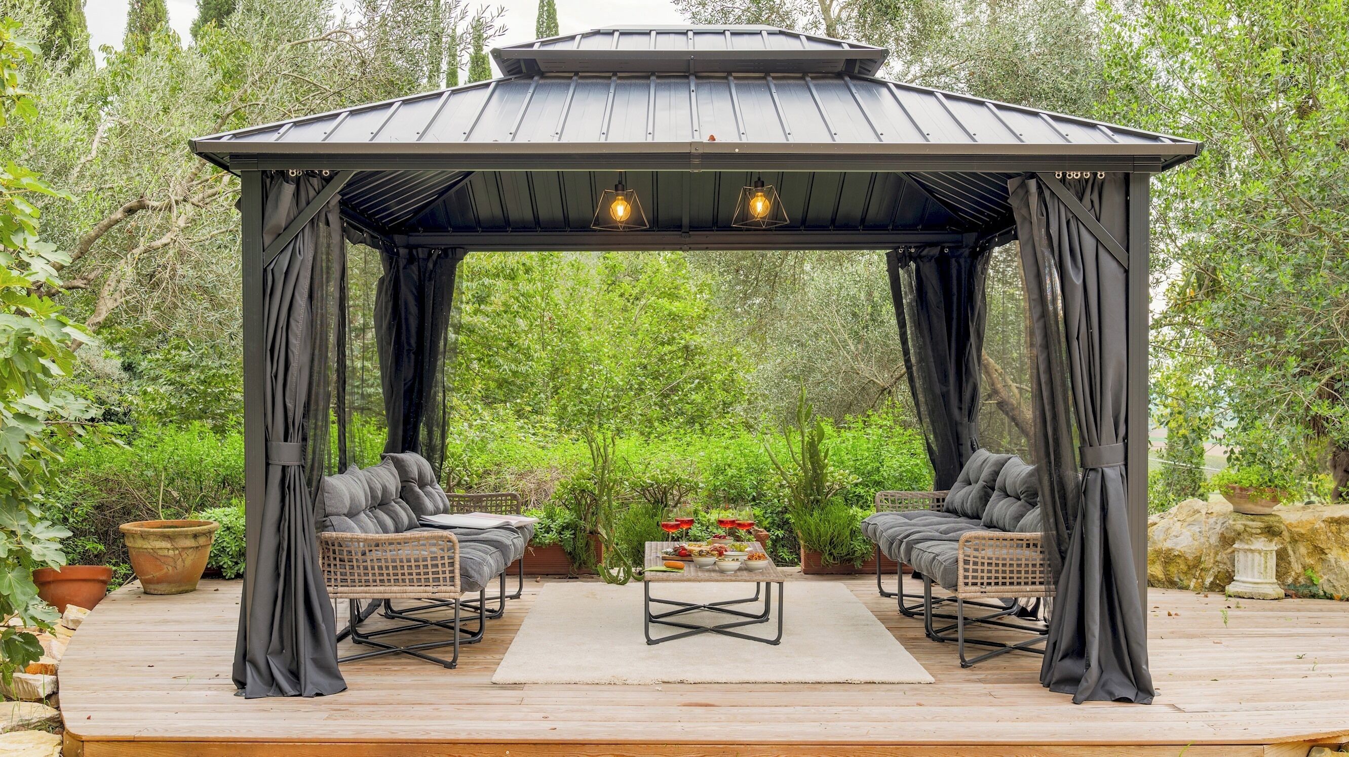 A gazebo with two cushioned chairs and a table set for dining, surrounded by lush greenery.