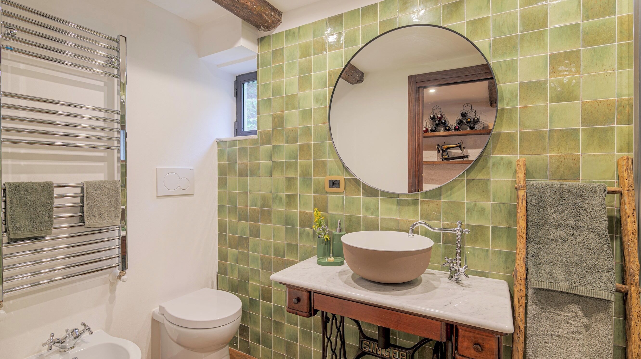 A bathroom with a white toilet, a round mirror, a sink with a beige basin, and a towel rack with two towels.