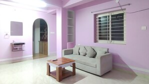 A living room with a white sofa, a wooden coffee table, a ceiling fan, a window with blinds, and a pink wall. - The Lakeview Classic Homestay (Agartala)