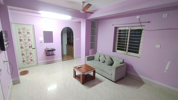 A living room with a sofa, a coffee table, a ceiling fan, a window with blinds, and a door leading to another room. - The Lakeview Classic Homestay (Agartala)