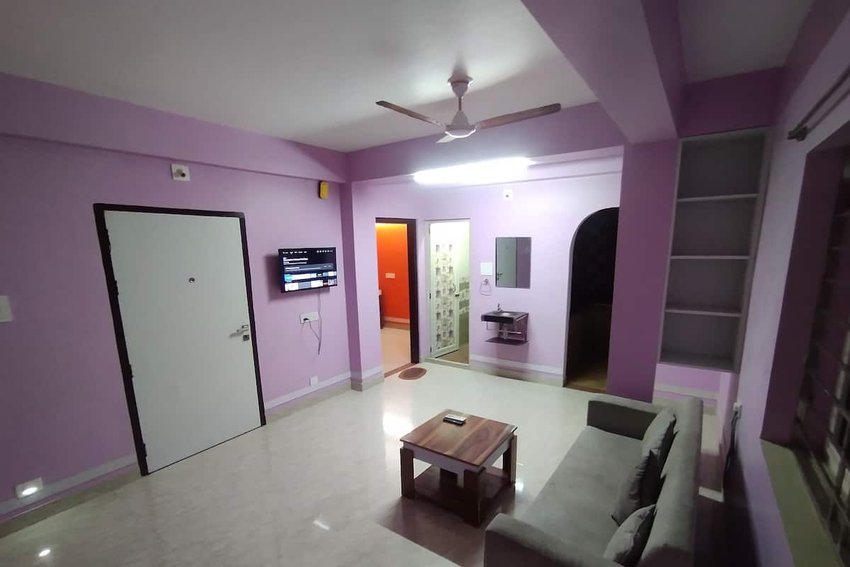 A room with a white door, a television mounted on the wall, a ceiling fan, a small table, and a sofa.