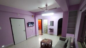 A room with a white door, a television mounted on the wall, a ceiling fan, a small table, and a sofa. - The Lakeview Classic Homestay (Agartala)