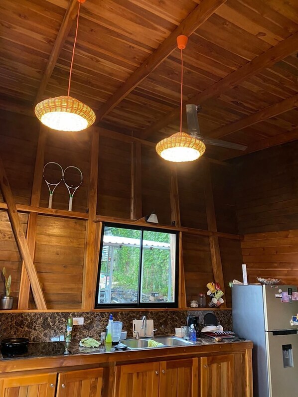 A rustic kitchen with wooden beams, a window with a view, and two hanging pendant lights. - Hoja Azul (Hojancha)