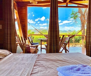 A wooden cabin room with a bed, a hammock, and a view of a pool and mountains. - Hoja Azul (Hojancha)