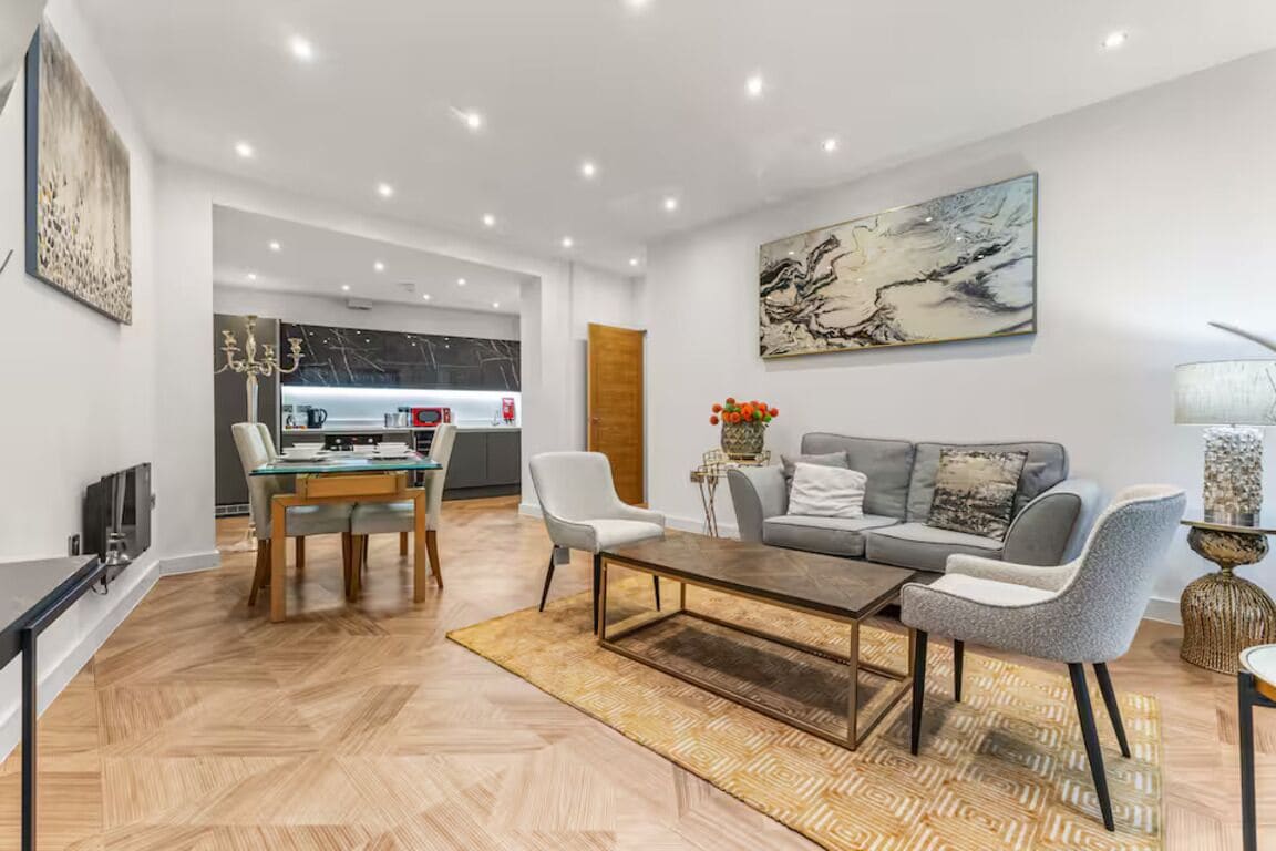 Un salon moderne avec un canapé, une table basse et un téléviseur. Il y a également un coin repas avec une table et des chaises. La pièce est dotée d’un parquet et d’un grand tableau abstrait accroché au mur.