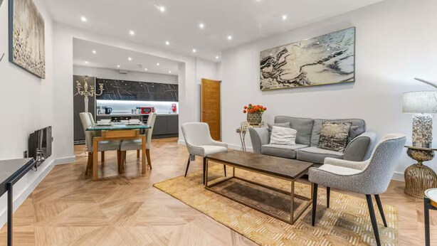 A modern living room with a sofa, coffee table, and TV. There is a dining area with a table and chairs. The room has wooden flooring and a large abstract painting on the wall. - Mill Conversion Flat Mins from Printworks - Pass the Keys (Salford)
