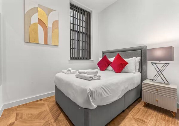 Un dormitorio moderno con cama gris, ropa de cama blanca y almohadas rojas. Se ve una ventana con persianas verticales, una mesita de noche con lámpara y una obra de arte en la pared.