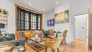 A modern living room with a sofa, coffee table, and chairs, a dining area with a table and chairs, a large window with blinds, and a painting on the wall. - Beautifully 3Bed Restored Apartment near Deansgate - Pass the Keys (Salford)