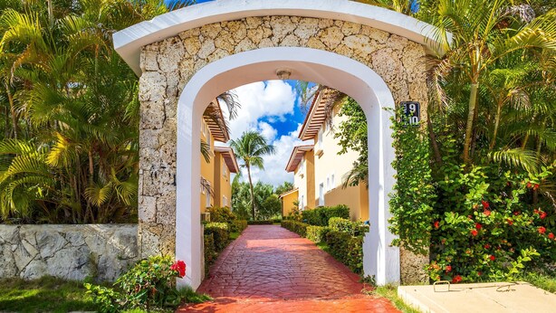 Un sendero atraviesa un arco de piedra que lleva a un edificio con fachada blanca y techo de tejas rojas, rodeado de exuberante vegetación y palmeras.