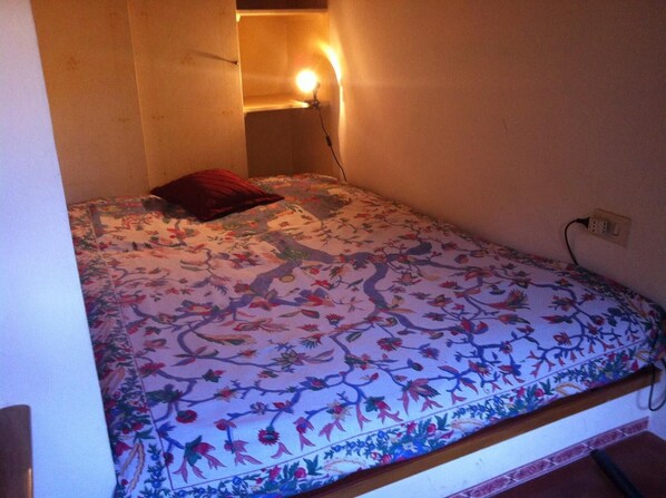 A small, dimly lit room with a single bed, a red pillow, and a wall-mounted light.