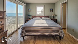 A bedroom with a large bed, a window with a view of the ocean, and a wooden floor. - Beachy McBeach House: Bold, Bright & Beachy (Surf City)