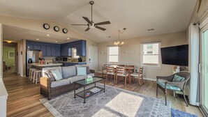 A spacious living area with a sofa, coffee table, and dining set. A kitchen with blue cabinets and a ceiling fan is visible in the background. - Beachy McBeach House: Bold, Bright & Beachy (Surf City)