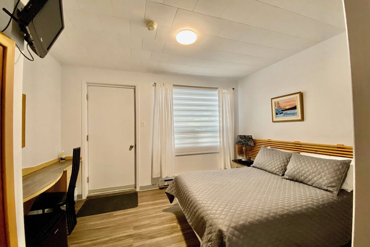 A hotel room with a bed, a desk, a chair, a television, a window with blinds, and a painting on the wall.