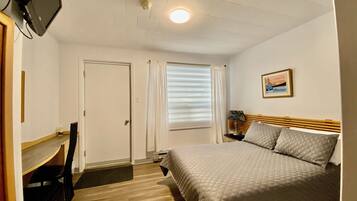 A hotel room with a bed, a desk, a chair, a television, a window with blinds, and a painting on the wall.