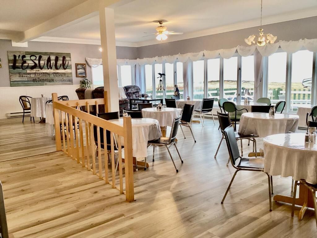 A spacious dining area with multiple round tables and chairs, a wooden staircase, and large windows offering a view of the outdoors.