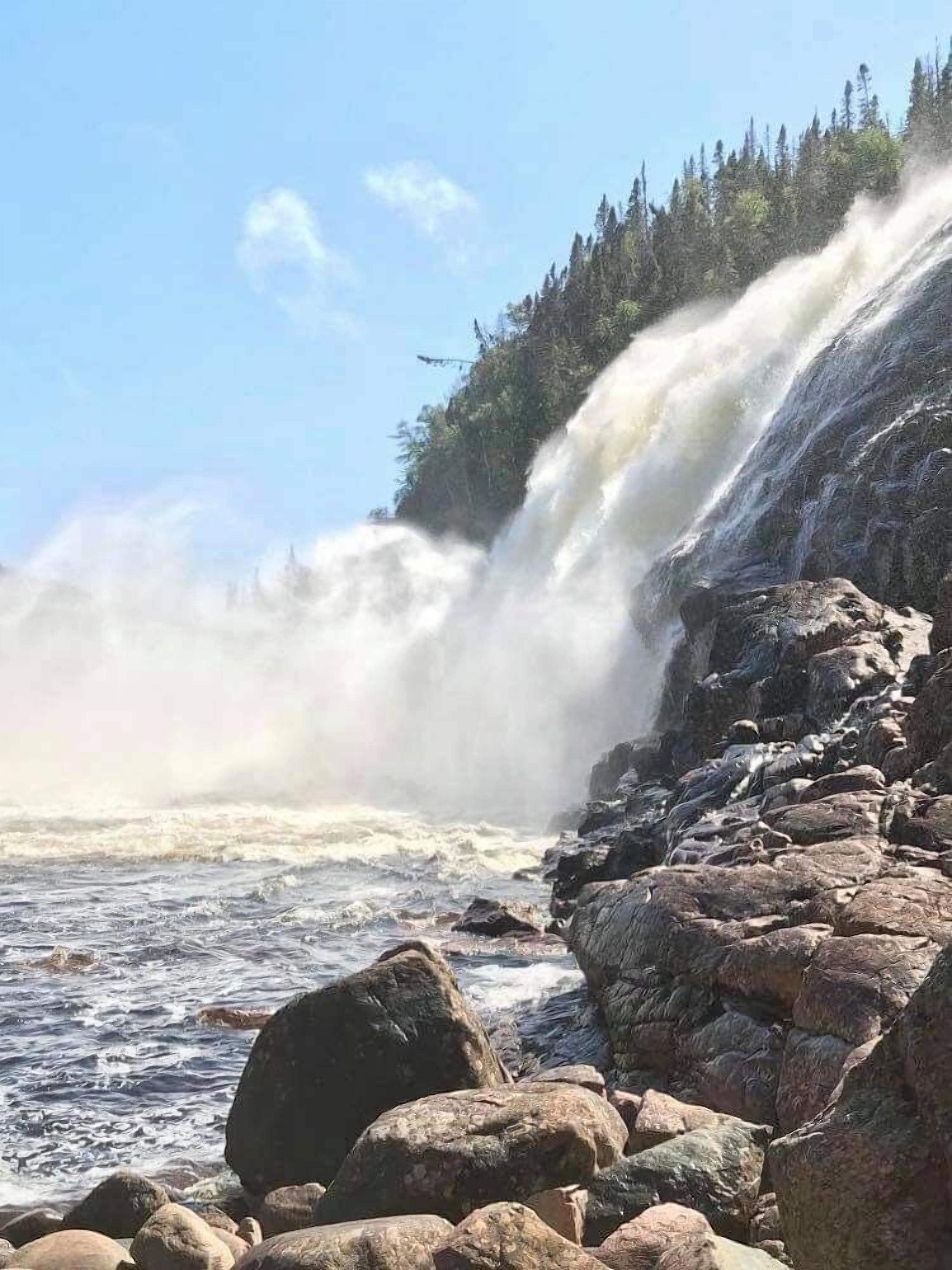 Ein Wasserfall stürzt über Felsen in ein Gewässer, umgeben von bewaldetem Gelände.