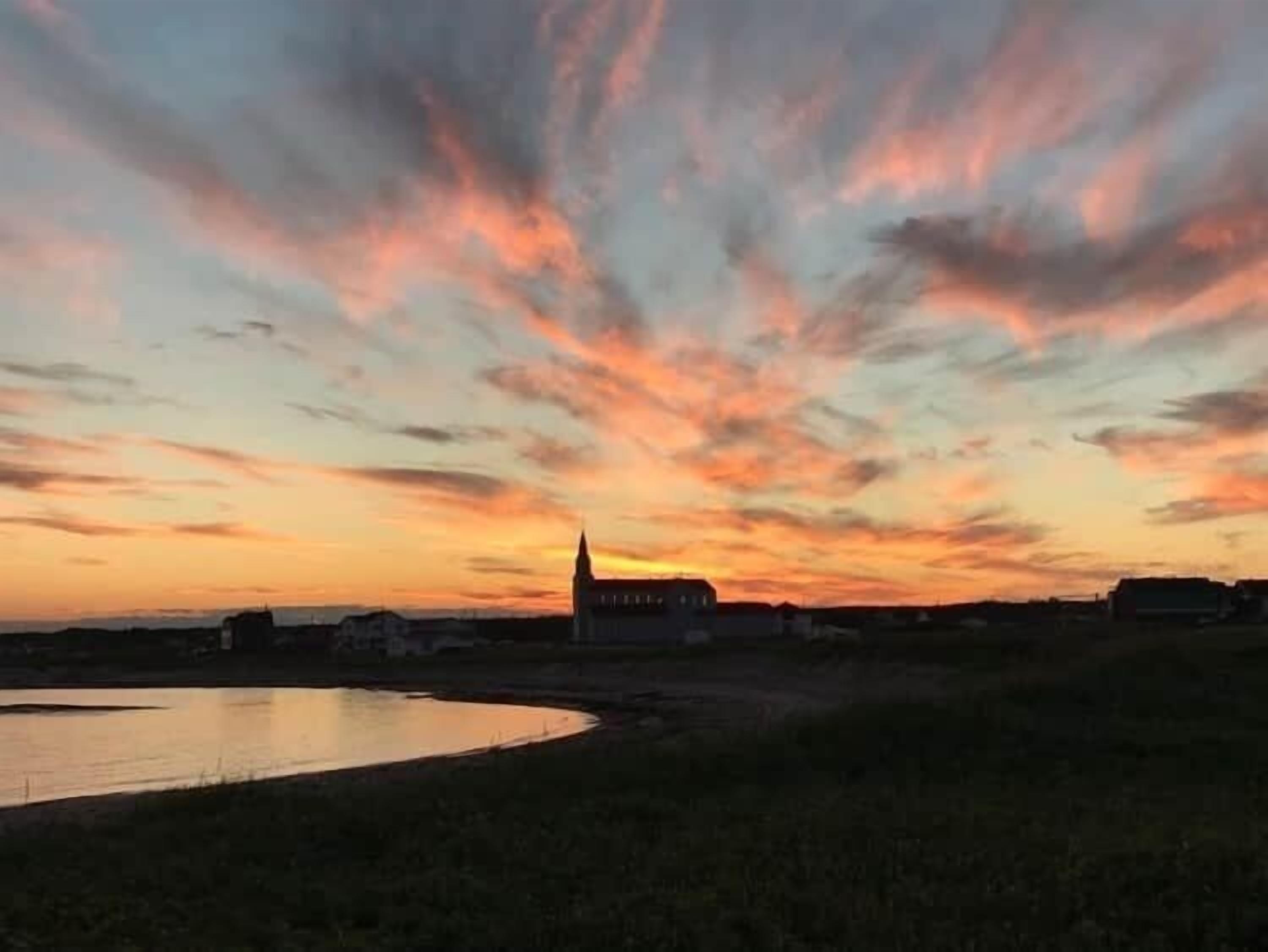 Un coucher de soleil sur une étendue d’eau, avec la silhouette d’une église et des bâtiments éloignés à l’horizon.