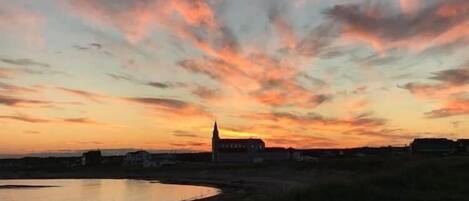 Un coucher de soleil sur une étendue d’eau, avec la silhouette d’une église et des bâtiments éloignés à l’horizon.