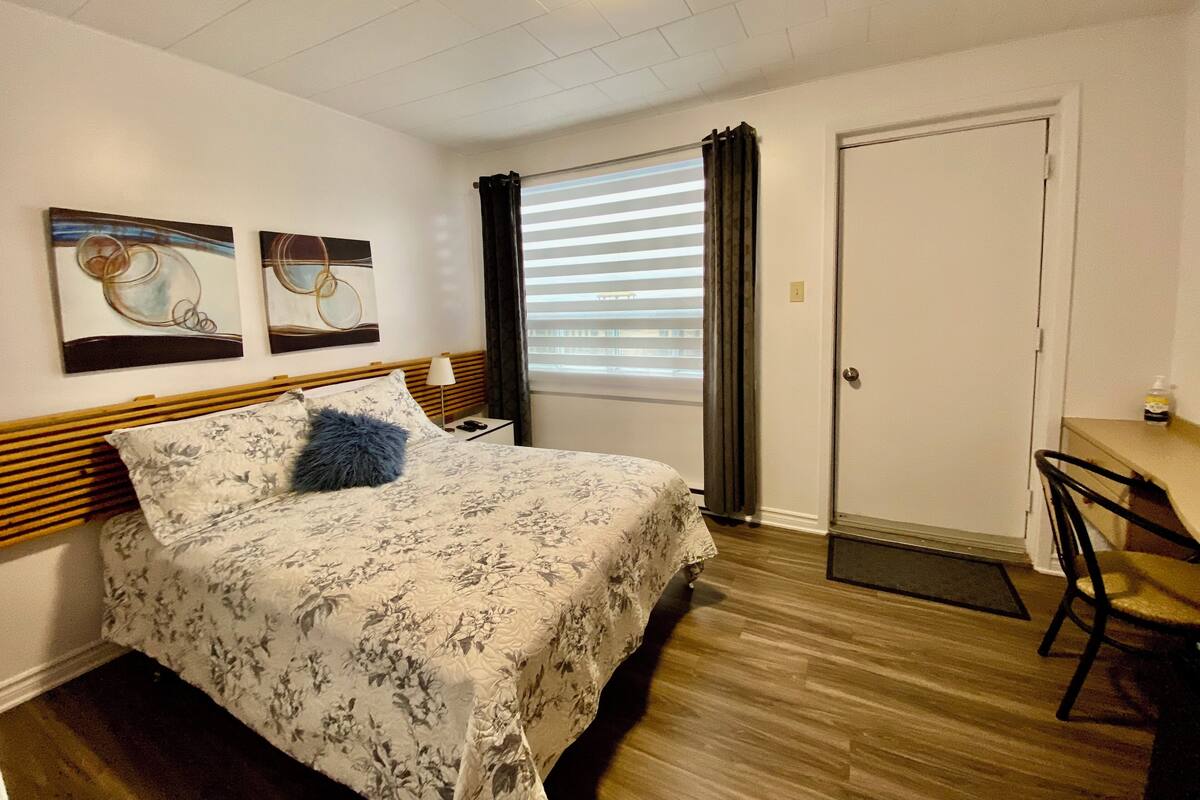 A bedroom with a bed, two wall art pieces, a window with blinds, a door, and a desk with a chair.