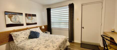 A bedroom with a bed, two wall art pieces, a window with blinds, a door, and a desk with a chair.