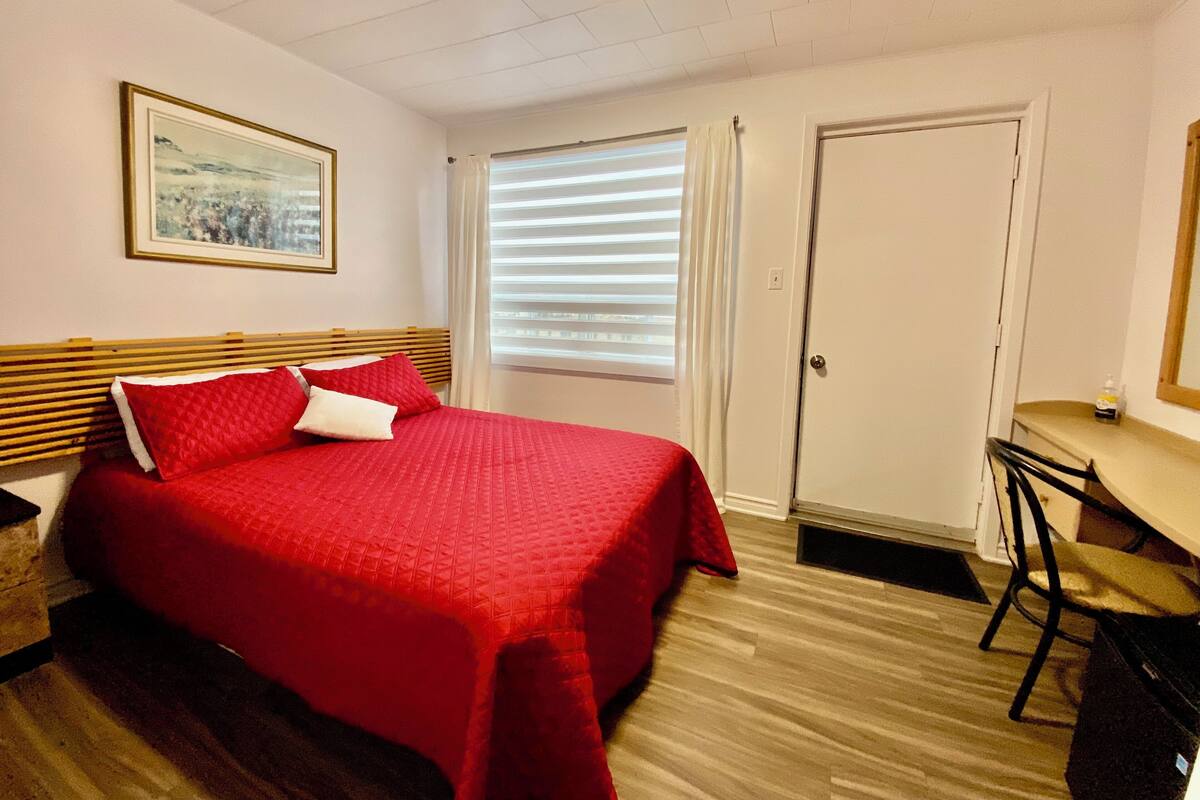 A bedroom with a bed, a desk, a chair, a door, and a window with blinds.