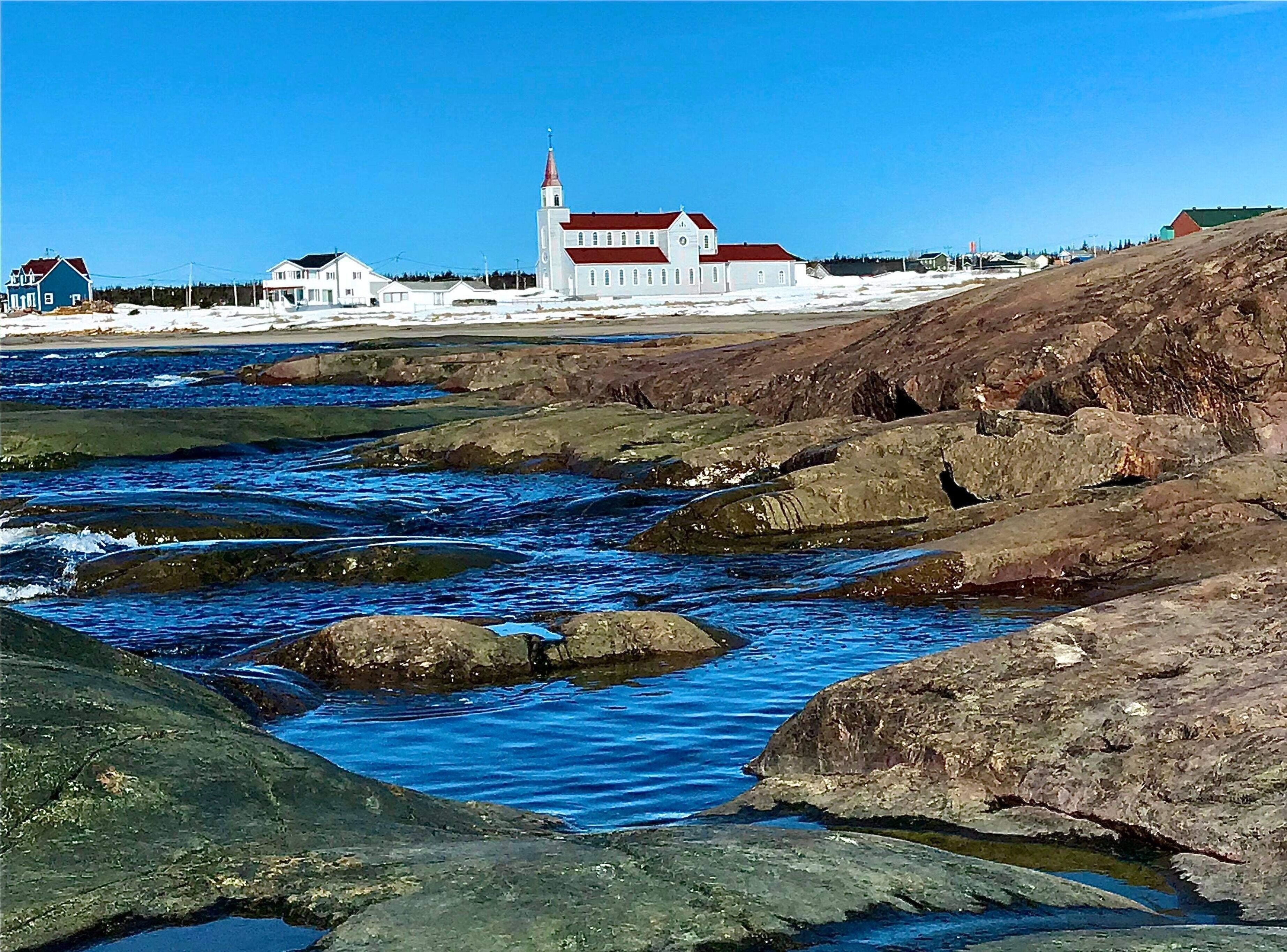 A coastal landscape with a white church with a red roof, rocky shoreline, and clear blue water.