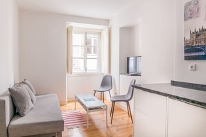 A compact living space with a sofa, two chairs, a small table, a TV, and a kitchen area with a countertop.