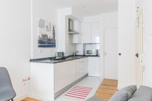 Cocina compacta con armarios blancos, microondas y una pequeña isla. Hay un cuadro de un paisaje urbano en la pared, y un extintor está montado allí mismo.