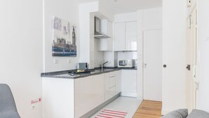 Cocina compacta con armarios blancos, microondas y una pequeña isla. Hay un cuadro de un paisaje urbano en la pared, y un extintor está montado allí mismo.