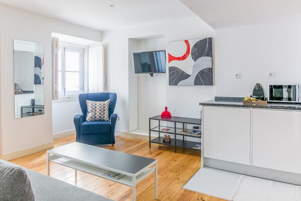 A modern living room with a blue armchair, a coffee table, a TV, and a kitchen area with a countertop and cabinets.