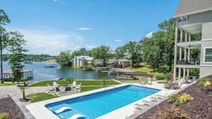 Piscina com vista para um lago, rodeada por um deck com móveis de exterior e área coberta.